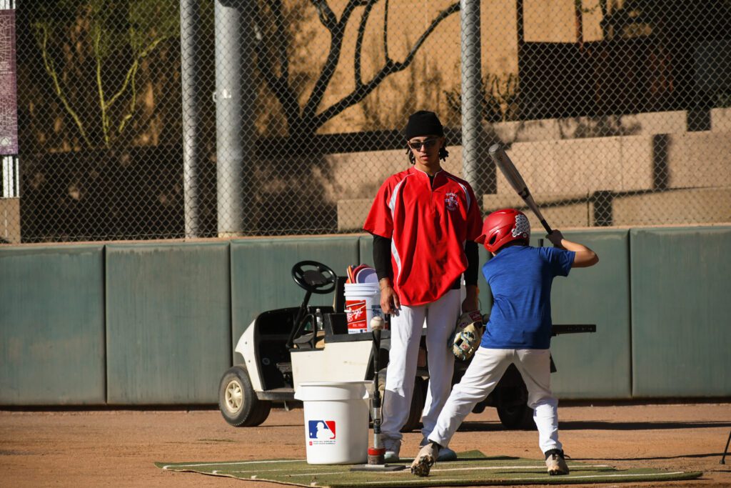 Young baseball player swings bat during practice with coach nearby.