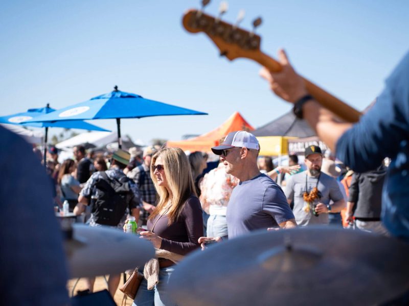 Outdoor crowd enjoying live music under blue sky and umbrellas.