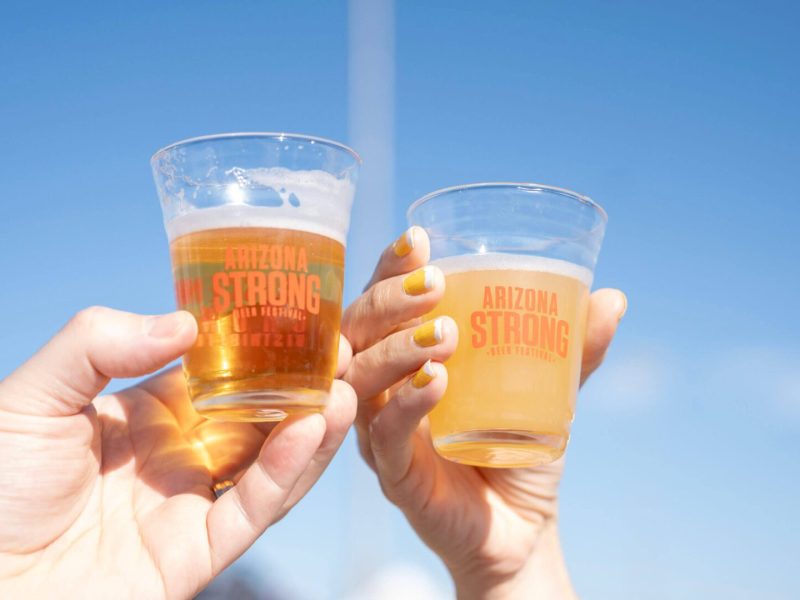 Two hands holding glasses of beer against a clear blue sky.