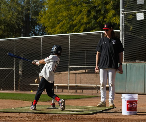 Young baseball player swings bat while coach watches on.